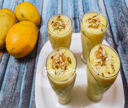 Mango milkshake by CookingPride served in 4 glasses on table, decorated by keeping 4 ripened mangoes besides the glasses